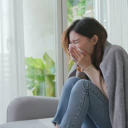 Woman holding her nose with a tissue while sitting on a couch.