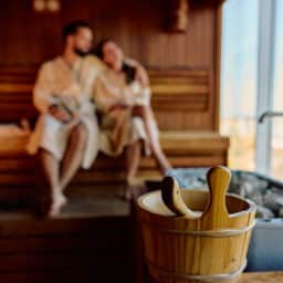 Blurred couple wearing bathrobes relaxing in a wooden sauna, enjoying the warmth and steam from the heater, with a bucket and scoop in the foreground.