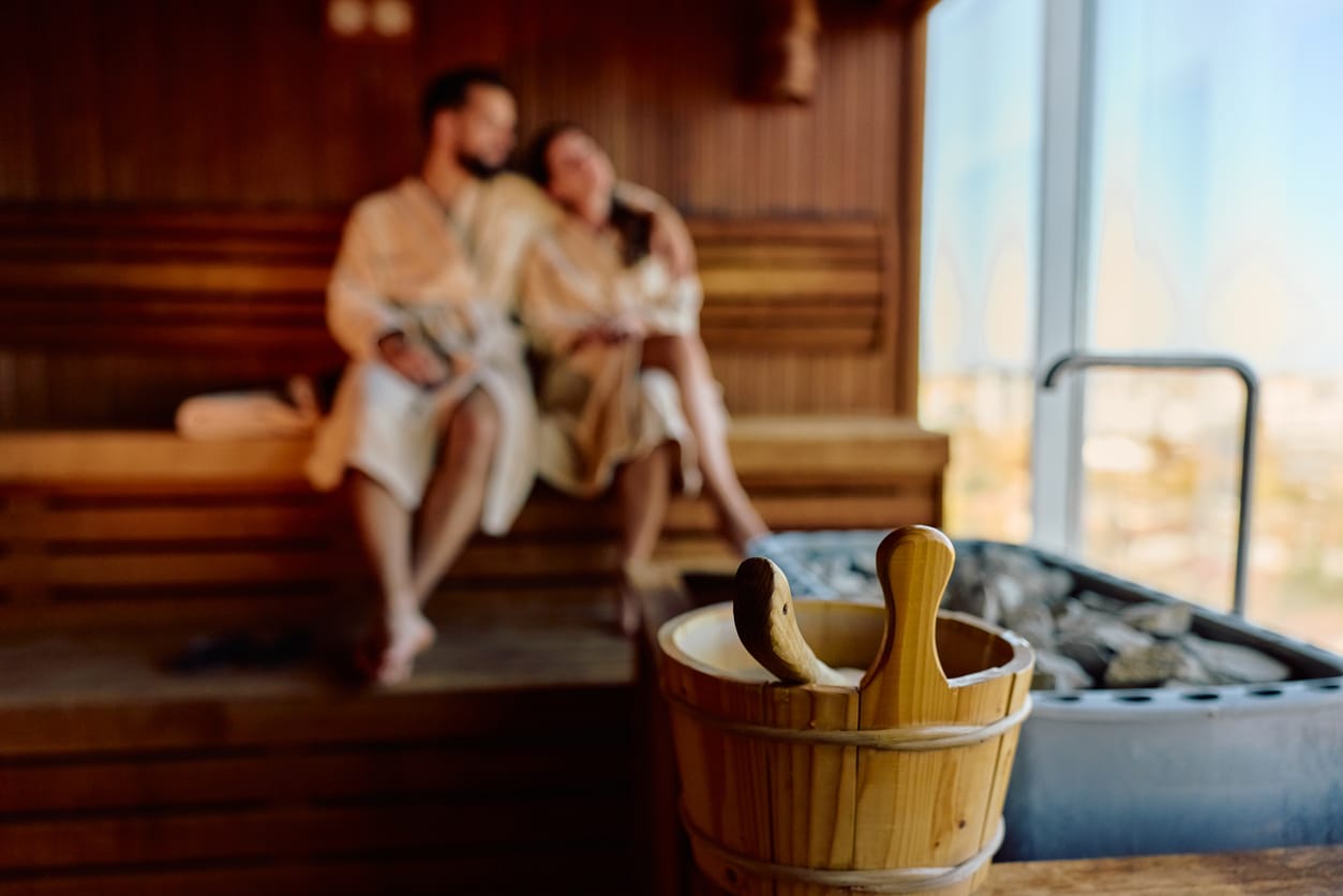 Blurred couple wearing bathrobes relaxing in a wooden sauna, enjoying the warmth and steam from the heater, with a bucket and scoop in the foreground.