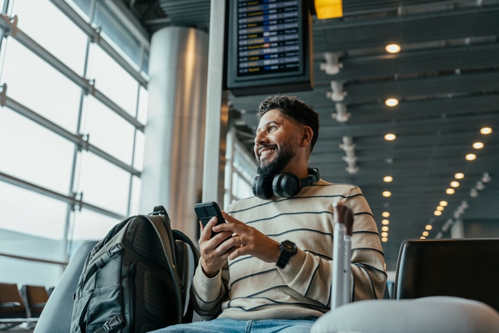 A man holding his smartphone and smiling while waiting to board at the airport.